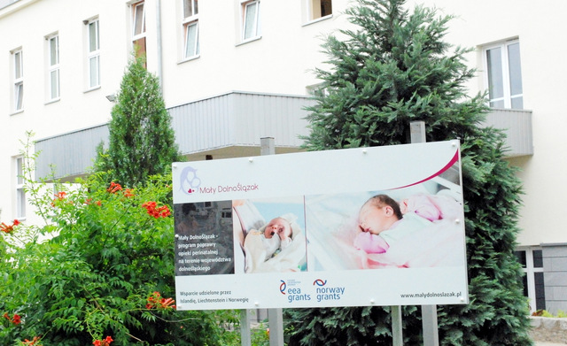 Przed szpitalem stoi tablica informująca o realizacji programu Mały Dolnoślązak zapraszająca do skorzystania z usług nowocześnie wyposażonych - bloku porodowego i oddziału neon